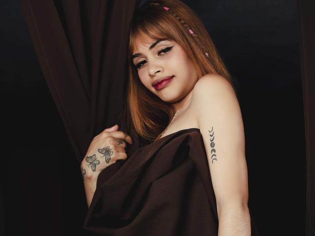Zenda Evans, avec ses cheveux bruns clairs, son rouge à lèvres rouge et ses tatouages sur le bras et la main, tient une robe marron, et regarde l'appareil photo avec un léger sourire. Fond noir..