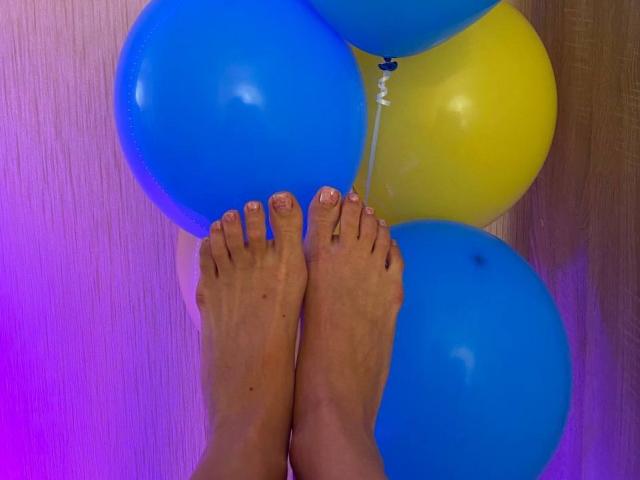 Les pieds nus d'AlicePrings, avec ses ongles peints, sont posés sur des ballons bleus et jaunes. Ses orteils sont légèrement recourbés, et les ballons sont regroupés..