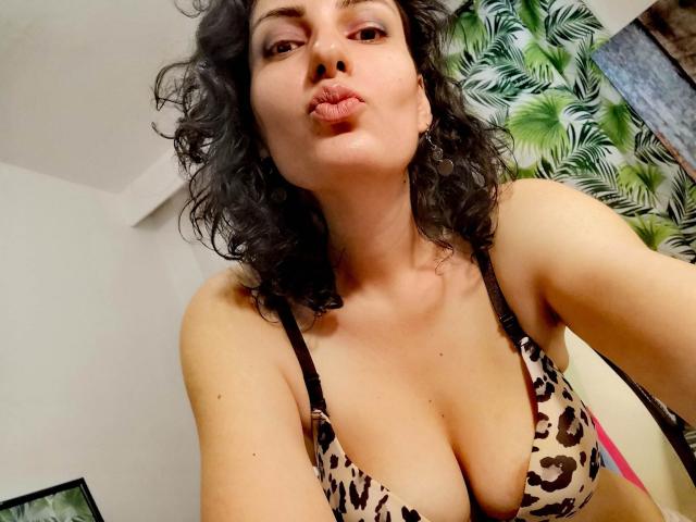 AddictedToUTS with curly dark hair, pouty lips, and a leopard print bra showing her tits, looking down at the camera, in a slightly messy room.