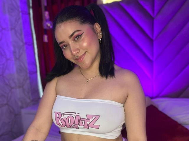 VeronicaWins, con due codini neri, eyeliner rosa e un top bianco che mostra la scritta "BOATZ" in rosa. Ha un sorriso carino, un seno piccolo e una collana con una croce dorata..