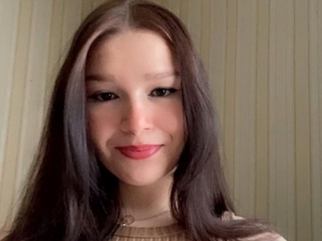 AvaCandy a de longs cheveux bruns, un eye-liner noir et du rouge à lèvres. Elle sourit avec une jolie figure et porte un haut beige. Ses seins ne sont pas visibles..