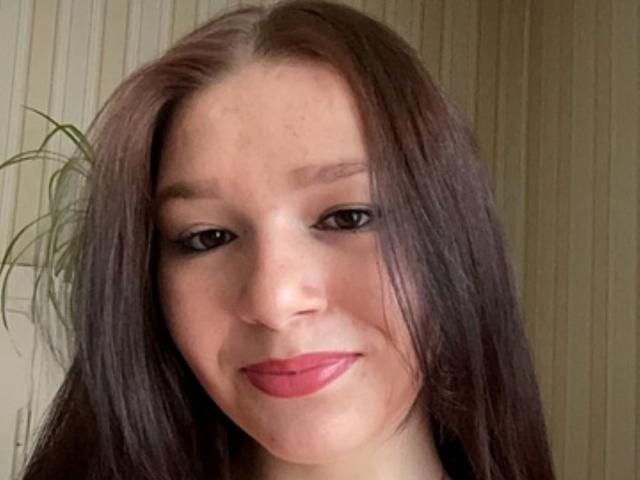 AvaCandy has long brown hair, red lipstick, and a slight smile. Her freckled face and black eyeliner stand out. She's looking down with a subtle, sexy expression.