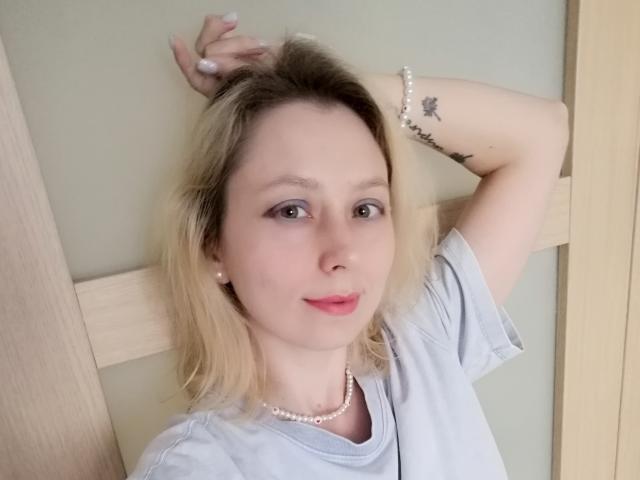 NaozuQAce con cabello rubio, piel clara, senos pequeños, collar de perlas, camisa blanca, tatuaje en el brazo, labios rosados y aretes de perlas, apoyada contra un marco de madera, con la mano en el cabello, sonriendo..