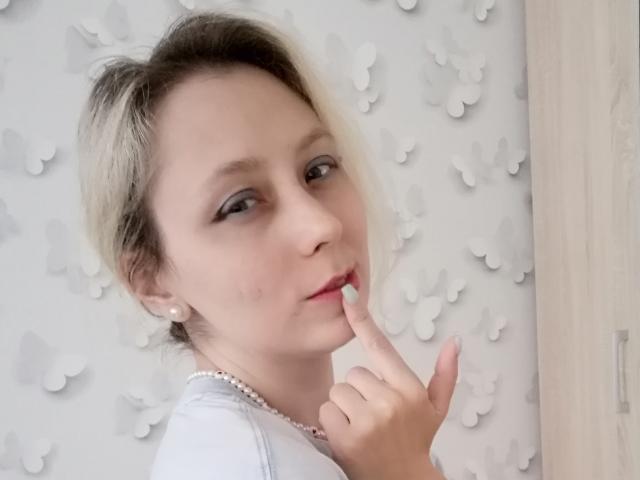 NaozuQAce tiene el cabello rubio claro, piel pálida y está chupándose el dedo mientras lleva un collar de perlas, vistiendo una camisa blanca, mostrando una expresión linda y ligeramente altanera..