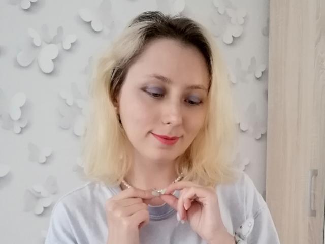 Eine Frau mit blonden Haaren, lila Lidschatten, pinkem Lippenstift und einer Halskette, die nach unten schaut und die Halskette berührt, trägt ein weißes Hemd und lächelt..