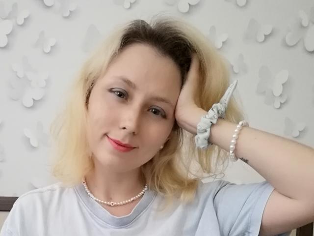 NaozuQAce tem cabelo loiro, pele clara e veste uma camisa branca com um colar de pérolas e um bracelete. Ela está tocando sua cabeça com um sorriso discreto..