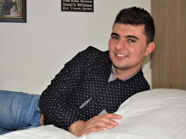 AndrethMuller, a cute guy with short dark hair, wearing a black polka dot shirt and blue jeans, lies on a white bed, smiling, showing his ass slightly.