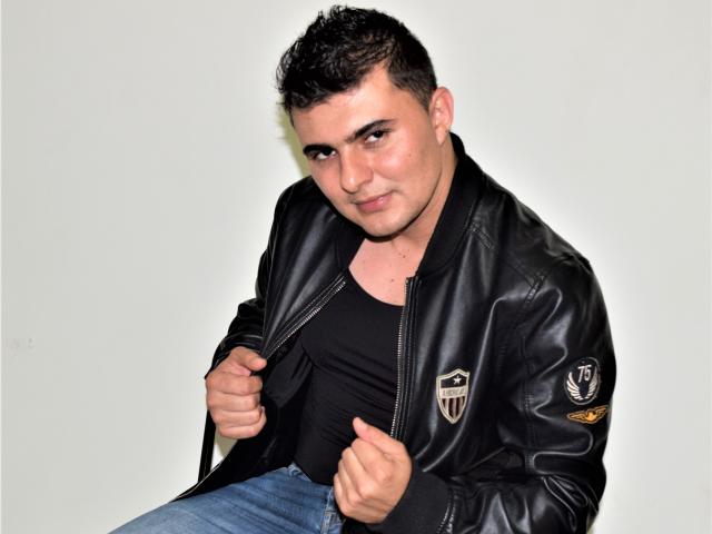 AndrethMuller, with short dark hair, black leather jacket, and black tank top, slightly pulling jacket, sitting, wearing blue jeans, looking at camera with a subtle smirk.