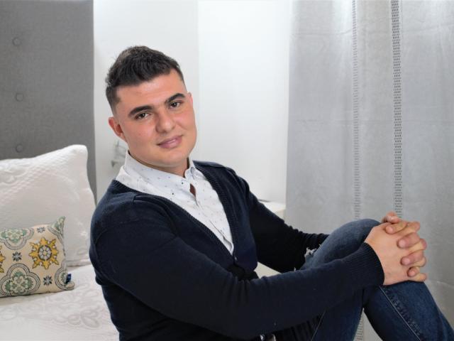 AndrethMuller has short black hair, a light skin tone, and is wearing a dark blue cardigan over a white shirt, sitting with hands on knees, looking at camera.