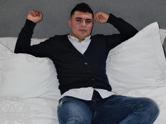 AndrethMuller, chubby dude, black cardigan, white shirt, blue jeans, arms raised, sitting on white bed with patterned pillows, short dark hair, slightly smiling, casual, relaxed.