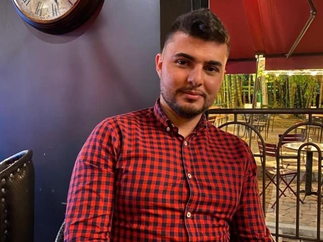 AndrethMuller with short dark hair, beard, and slight smile, wearing a red and black checkered button-up shirt, sitting indoors. Clock on blue wall. Red canopy outside.