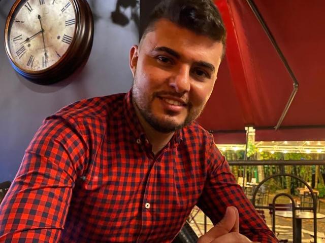 AndrethMuller, with a short beard and dark hair, wearing a red and black checkered shirt, smiling and giving a thumbs-up, clock on the wall behind him.