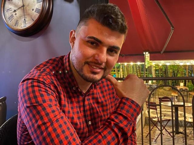 AndrethMuller, with a red and black checkered shirt, has a beard, short curly hair, and a slight smile, resting his chin on his hand. Clock in background.