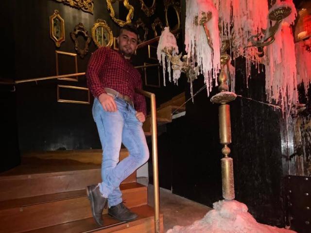 AndrethMuller, standing on wooden stairs, wearing a red plaid shirt, blue jeans, and gray boots, with a black railing and ice decorations in a dark, ornate room.