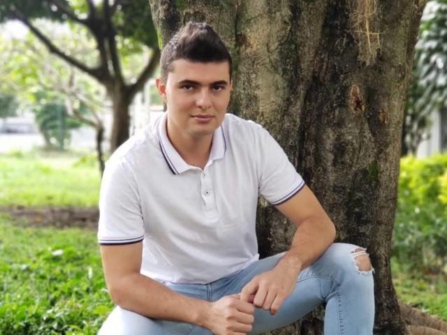 AndrethMuller sitting against a tree, wearing a white polo shirt with blue trim and ripped blue jeans, looking at the camera with a slight smile.