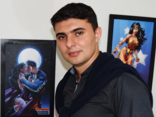 A photo of a handsome, dark-haired man with a black shirt and blue scarf, standing between two framed pictures of AndrethMuller as Wonder Woman and Superman kissing.