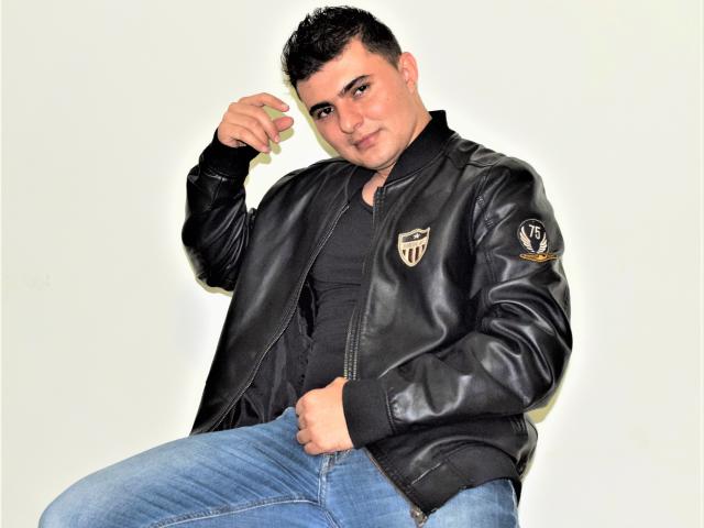 AndrethMuller with short black hair, black leather jacket, black shirt, and blue jeans, holding a green object near his head, sitting with a slight smirk, looking confident.
