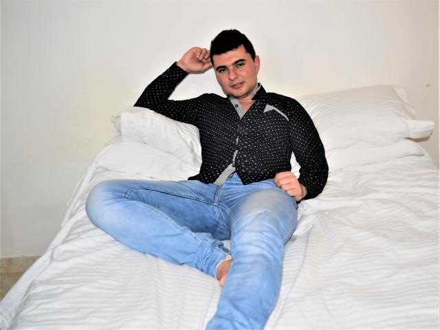 AndrethMuller, with short black hair, wearing a black polka dot shirt and ripped blue jeans, lies on a white bed, hand behind head, looking at camera.