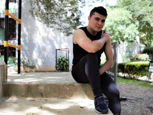 AndrethMuller, muscular, black sleeveless shirt, black leggings, grey sneakers, short black hair, sitting on concrete steps, arms crossed, smirking, daytime, outdoor, trees, greenery in background.