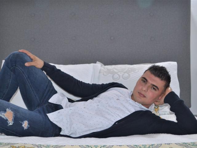 AndrethMuller is lying on a bed, wearing a white polka-dot shirt, blue jeans with rips, and a black jacket. He's looking at the camera with a relaxed expression.