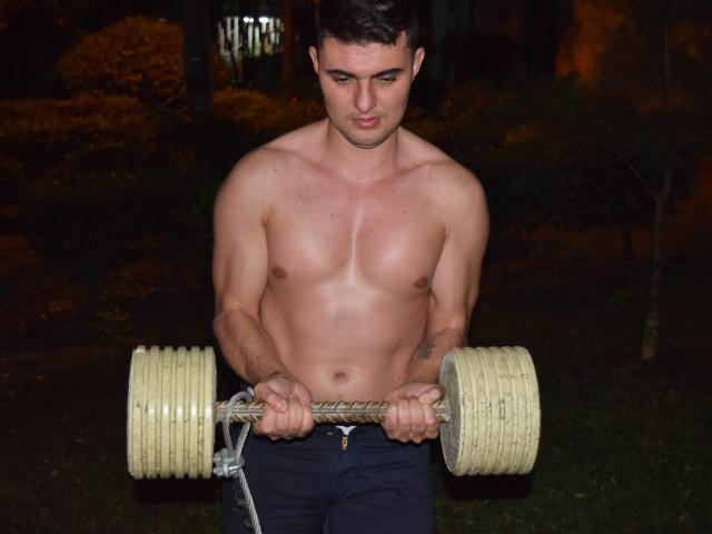 AndrethMuller is shirtless, holding a heavy dumbbell with both hands, looking down with a serious expression. He has a muscular, defined chest and is wearing dark pants.