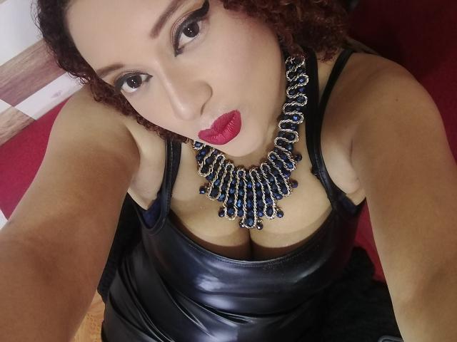 VioletaSwitch with curly brown hair, red lipstick, and black leather dress, wearing a blue and silver statement necklace, showing her boobs in a selfie.