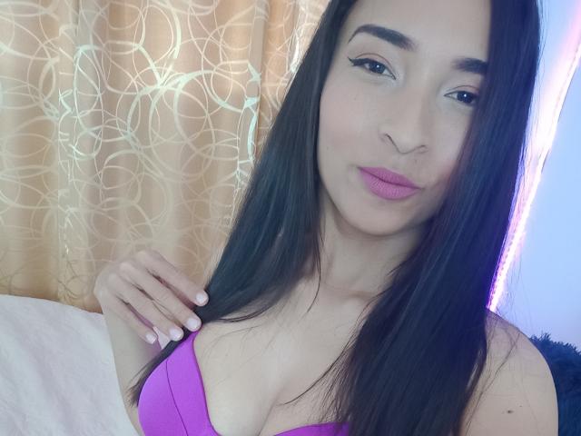 SarahyJhones with long black hair, pink lipstick, and a purple bra, showing a bit of her tits, holding her hair, smiling gently, in a bright indoor setting.