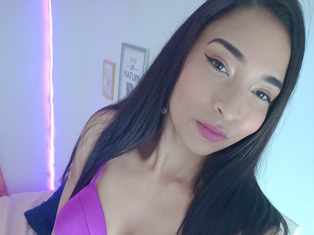SarahyJhones, con lunghi capelli neri, rossetto rosa e reggiseno viola, che mostra i suoi piccoli seni, e un'aria sottile e sensuale. Ha l'eyeliner applicato e un'espressione carina e sicura di sé..