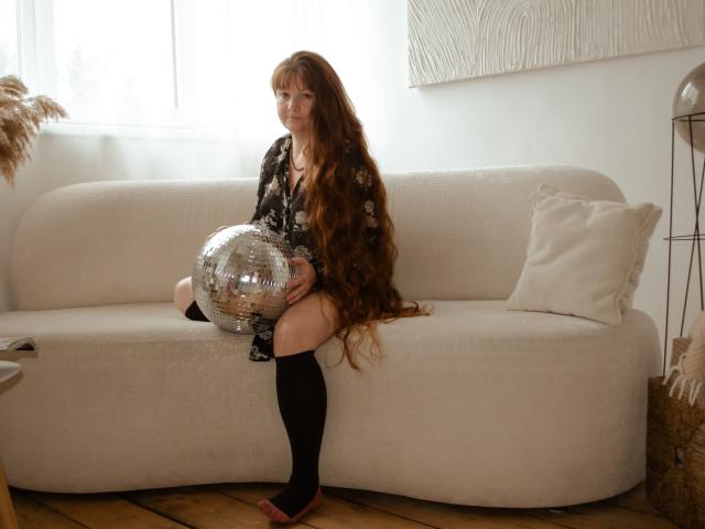 OVanessaHotly sits on a beige couch, holding a shiny silver disco ball, wearing a black floral dress, black knee socks, and red flats, with long red hair.