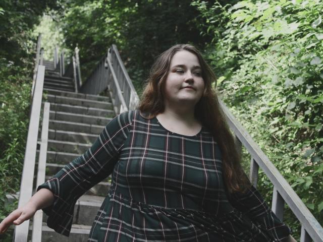 DaniellaGraceZZ has long brown hair, wears a green plaid dress, and stands on a stone staircase with metal railings, looking slightly to the side with a subtle smile.