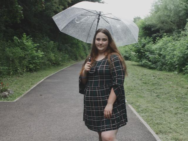 DaniellaGraceZZ with long brown hair, holding a transparent umbrella, wearing a green plaid dress, standing on a gray path, smiling, green foliage in background, overcast day.