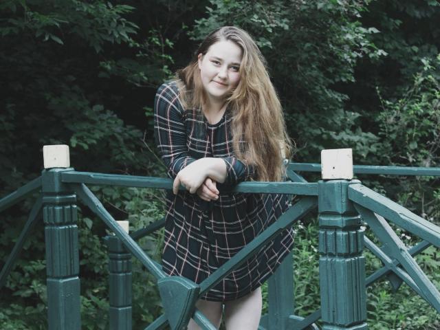 DaniellaGraceZZ with long brown hair, wearing a black plaid dress, leaning on a green wooden bridge, smiling, hands on railing, forest background.