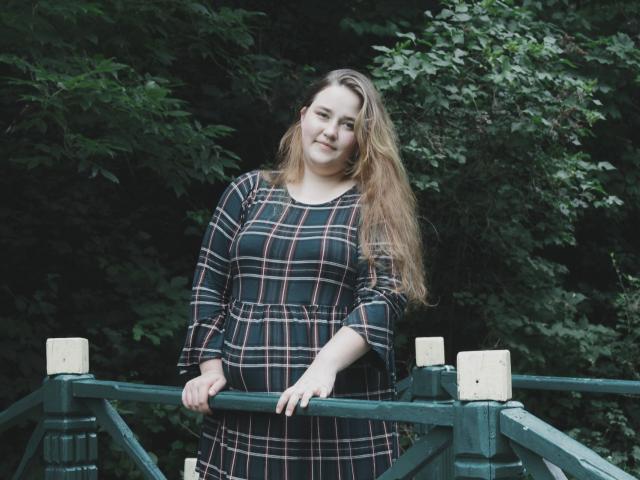 DaniellaGraceZZ with long brown hair, wearing a black plaid dress, standing with hands on green railing, smiling softly. Her tits are modest, and her pussy is not visible.