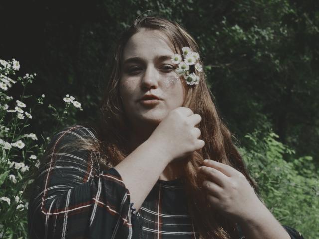 DaniellaGraceZZ, con cabello castaño, vestida con una camisa de cuadros, tiene flores blancas en el cabello, se ve ligeramente triste, sosteniendo su barbilla, en un bosque oscuro y verde..