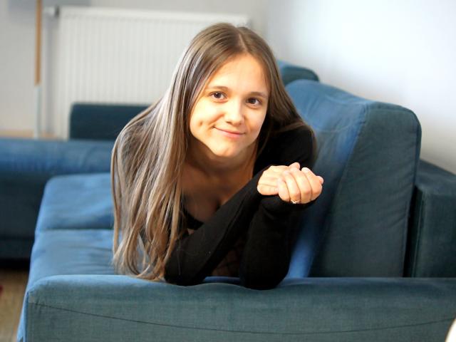Emily, met lang bruin haar, een zwarte top, leunend op een blauwe bank, lachend, met armen over elkaar, schattig, kleine borsten, bruine ogen, lichte huid, zachte belichting, casual, huiselijke setting..