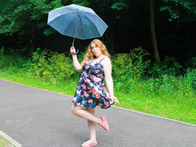 Jessika Daniels está de pie en una acera, sosteniendo un paraguas azul, vestida con un vestido floral, pantuflas rosas, con cabello rubio rizado, y con una figura curvilínea, de cadera mediana y senos pequeños..