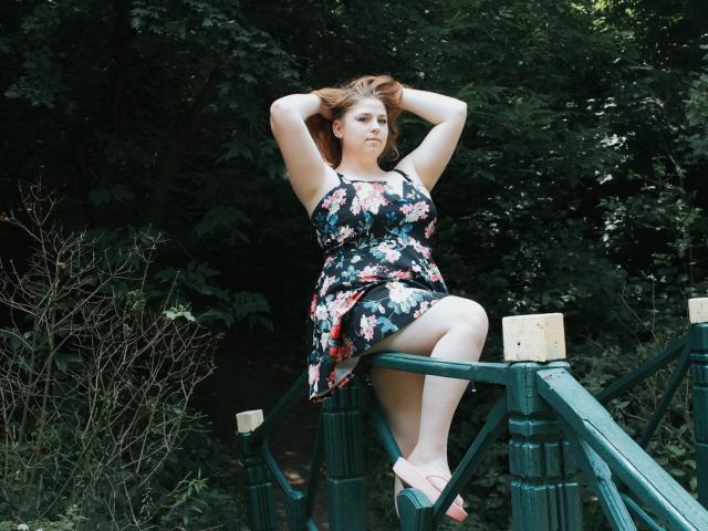 JessikaDaniels, met armen omhoog, zit op een groene houten brug, gekleed in een bloemenjurk, waarbij haar borsten en schaamte door de jurk te zien zijn, met rood haar en roze sandalen, op een groene achtergrond..