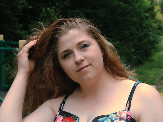 JessikaDaniels met golvend bruin haar, blauwe oogmake-up, een bh met bloemenprint, die lichtjes glimlacht, met haar hand in haar haar, in een buitenomgeving, met een groene achtergrond. Borsten zichtbaar, lichte huid, casual uitstraling..