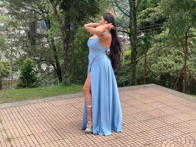 MiaSuarez with long dark hair, wearing a blue, floor-length dress with a high slit, showing her tits and ass, standing on a brick patio, touching her hair.