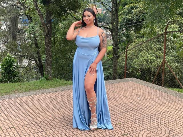 MiaSuarez with big tits, wearing a blue, thigh-high slit dress, and silver heels, standing on a brick patio, touching her hair, green tattoo on her left arm.