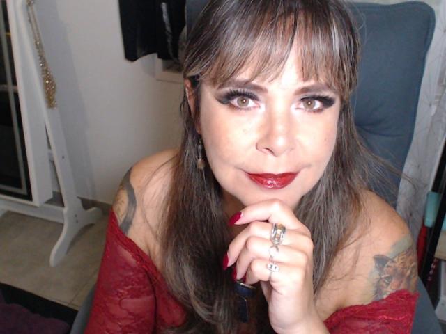 EvaLuv has brown hair, red lipstick, and dark eyeliner, wearing a red lace top, with tattoos on her shoulder, red nails, and rings, looking up with a slight smile.