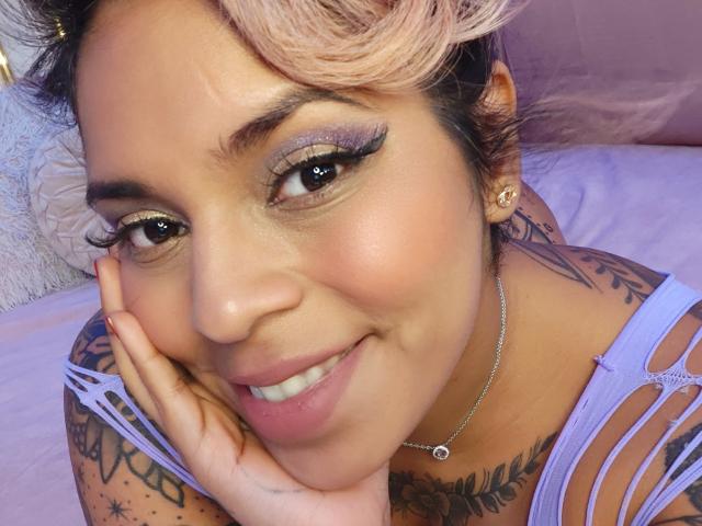 Chili, una milf heteroflexible de pelo rubio platino, sonríe mientras se toca la mejilla, mostrando su lencería blanca todos sus tatuajes.