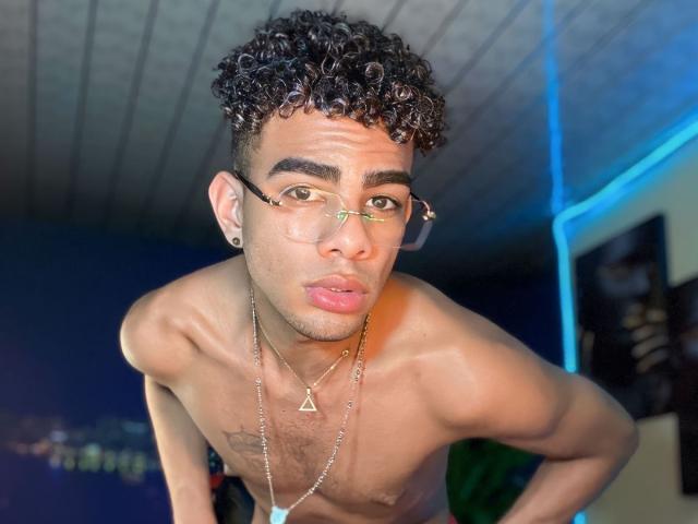 JamesDream, shirtless with curly black hair, glasses, and a triangular necklace, leaning forward with a pouty expression, showing off his toned chest and tattoos, has medium brown skin.