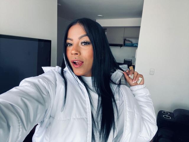 Amberv with long black hair, white puffy jacket, and tongue out, holding jacket collar, showing white ring on finger, looking at camera with slightly open mouth.