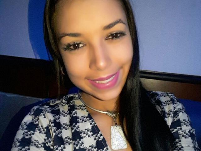 NicoleLoveGirl with dark straight hair, brown eyes, and pink lipstick, wearing a blue and white patterned top, smiling at the camera. She has a necklace with a white pendant.