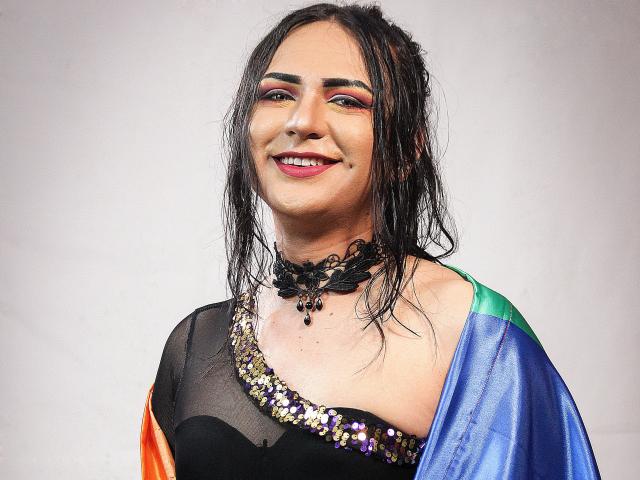 Iris Durand hat feuchtes, schwarzes Haar, auffälliges Make-up und eine schwarze Kette mit Perlen. Sie trägt ein glänzendes schwarzes Oberteil mit Pailletten und einen farbenfrohen Umhang mit Regenbogenstreifen. Iris Durand lächelt..