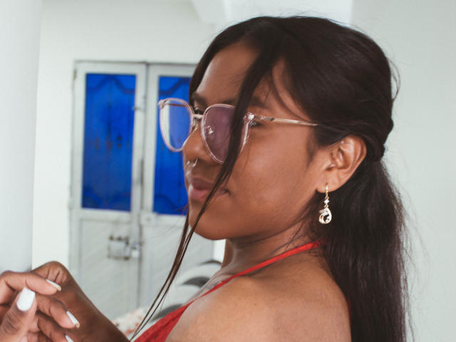 KimmySolange with dark brown skin, wearing pink glasses, red halter top, and dangling earrings, holding a white ring, smiling slightly, long black hair, nose ring.
