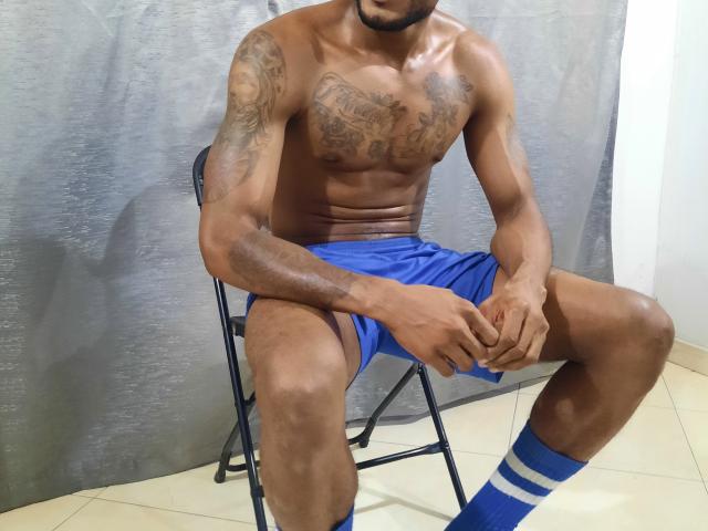 JeyKon, shirtless, with tattoos on chest and arm, wearing blue shorts, blue and white socks, sitting on black chair, hands on knees, in a room with tiled floor.