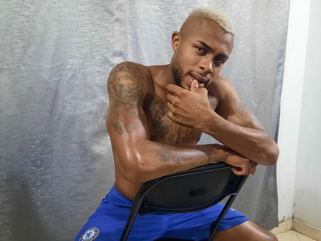 JeyKon, shirtless, muscular, tattooed, with bleached hair, blue shorts, biting finger, leaning on black chair, looking at camera, sweaty, in a room with a silver curtain.