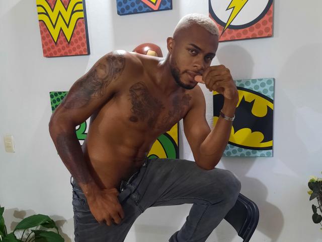 Shirtless JeyKon with tattoos, dark skin, and a shaved head, leans on one knee, wearing gray jeans, with a pensive expression, in a room with colorful comic art.